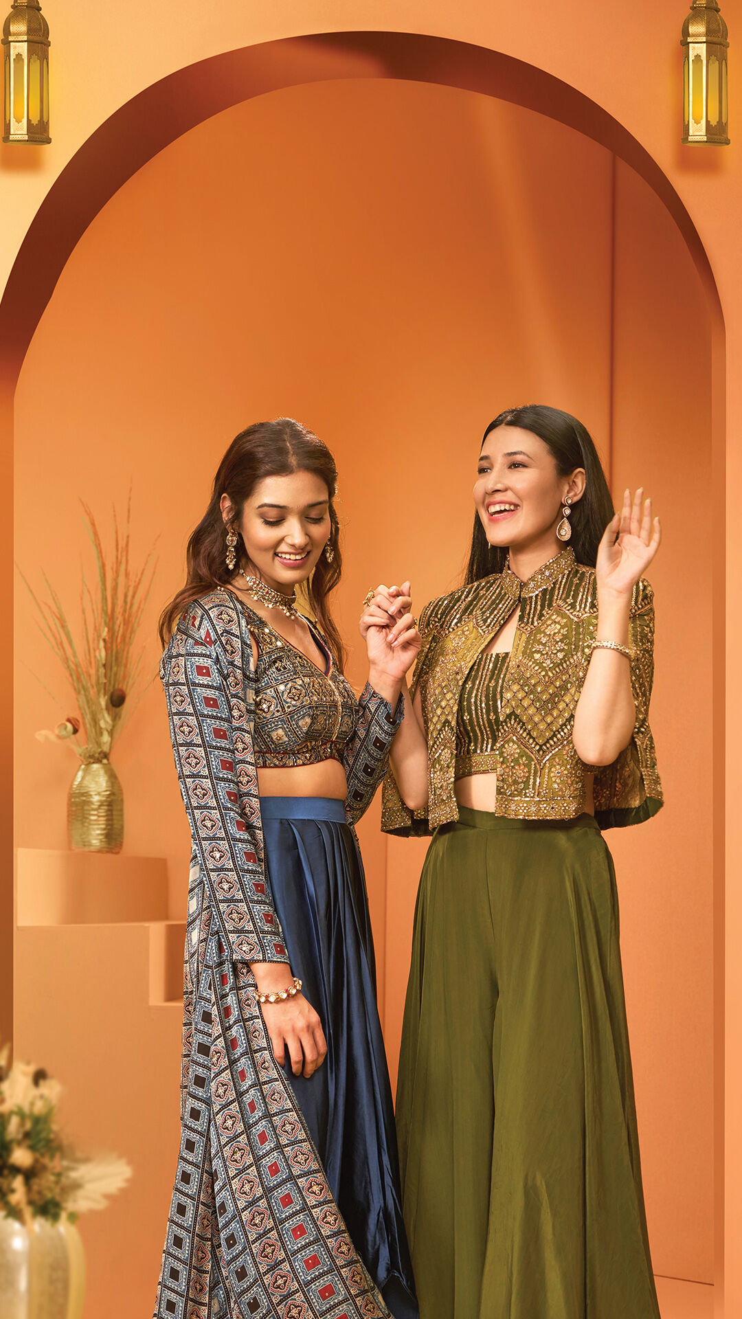 Two women in modern Indo-western outfits smiling under an archway with lanterns, dressed in a printed lehenga set and a stylish crop top with palazzo pants